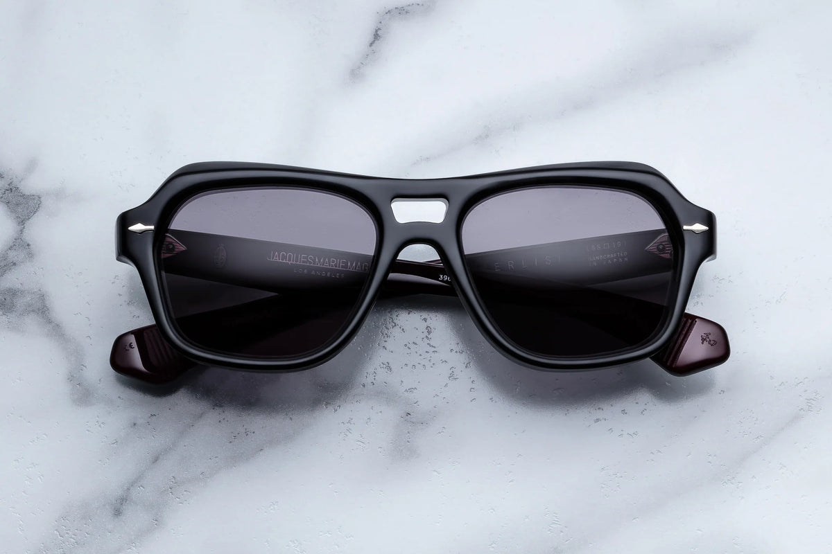 Jacques Marie Mage Tamerlis sunglasses from Jacques Marie Mage, featuring thick frames and dark lenses, rest on a white marble surface and showcase a daring silhouette with sculptural temples.