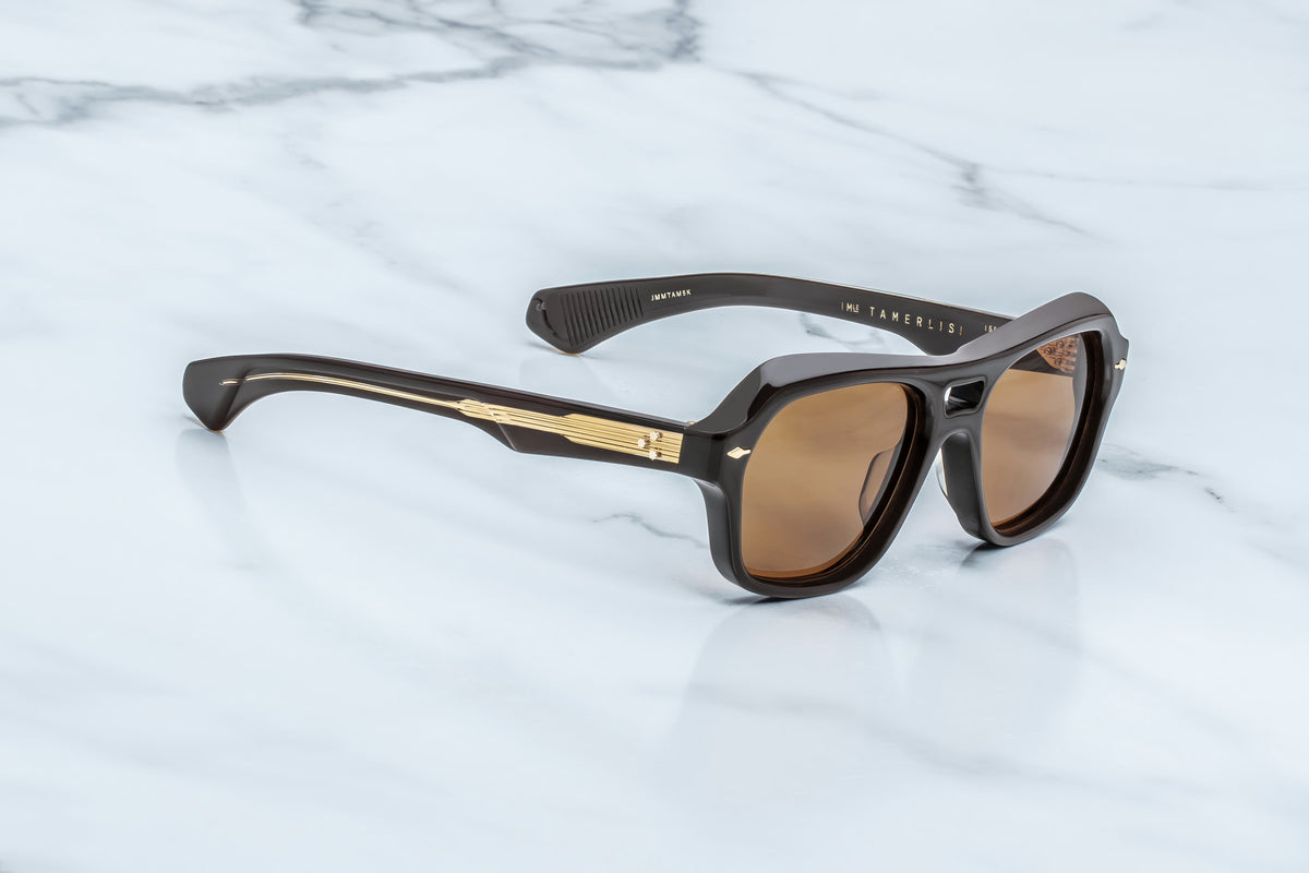 Jacques Marie Mage Tamerlis sunglasses by Jacques Marie Mage, featuring a daring silhouette with black frames, brown lenses, gold detailing on the arms, and sculptural temples, are displayed on a white marble surface.