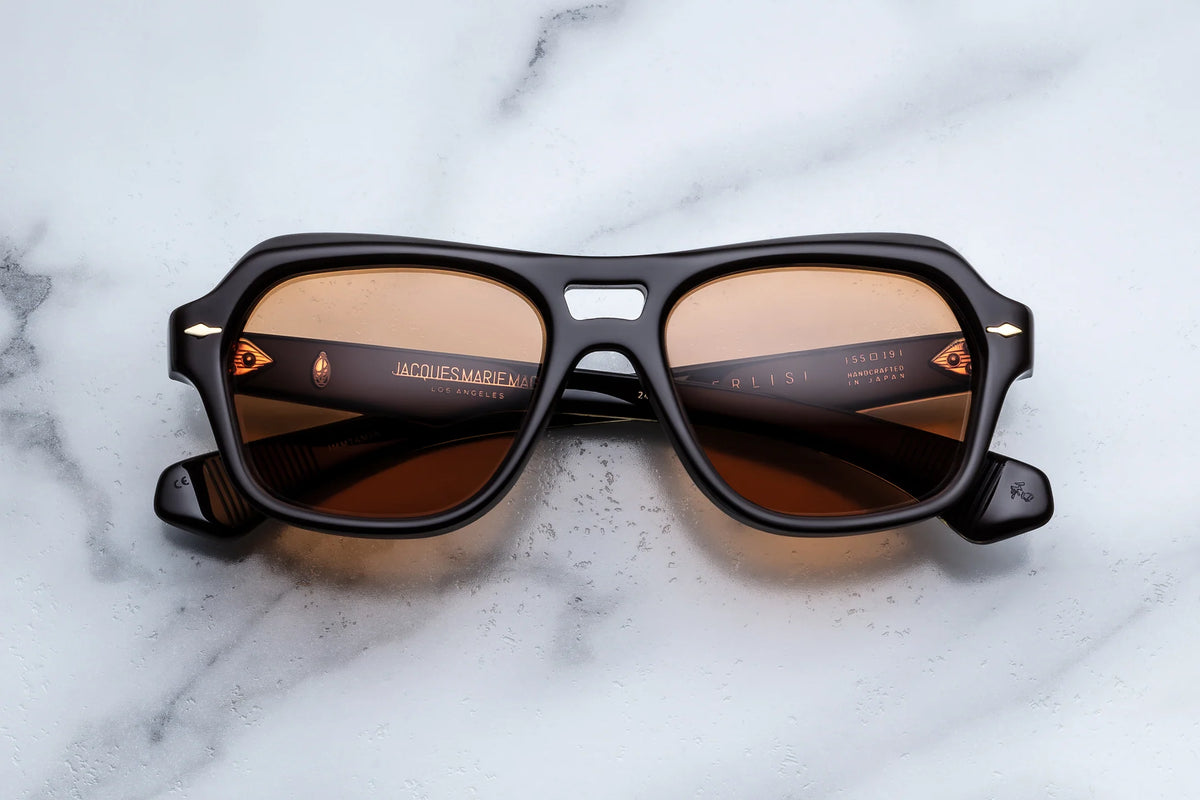 A pair of Jacques Marie Mage Tamerlis sunglasses in black, featuring rectangular frames with brown-tinted lenses, rests on a white marble surface. The sculptural temples enhance the sophisticated design, while gold text bearing the Jacques Marie Mage brand details adorns the arms for an elegant touch.