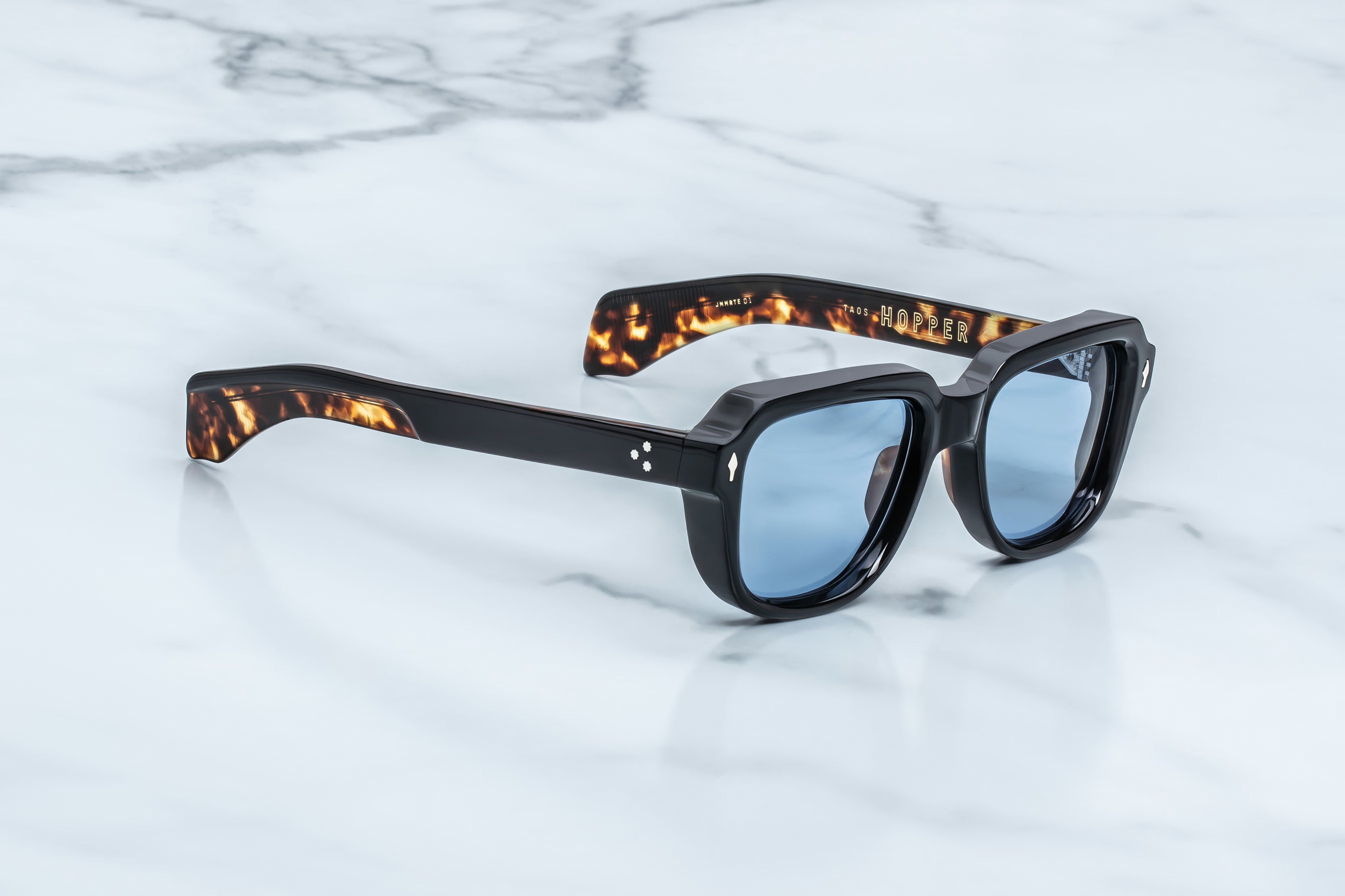 A pair of square-frame sunglasses with black frames, tortoiseshell-patterned temples, and light blue lenses, resting on a white marble surface.