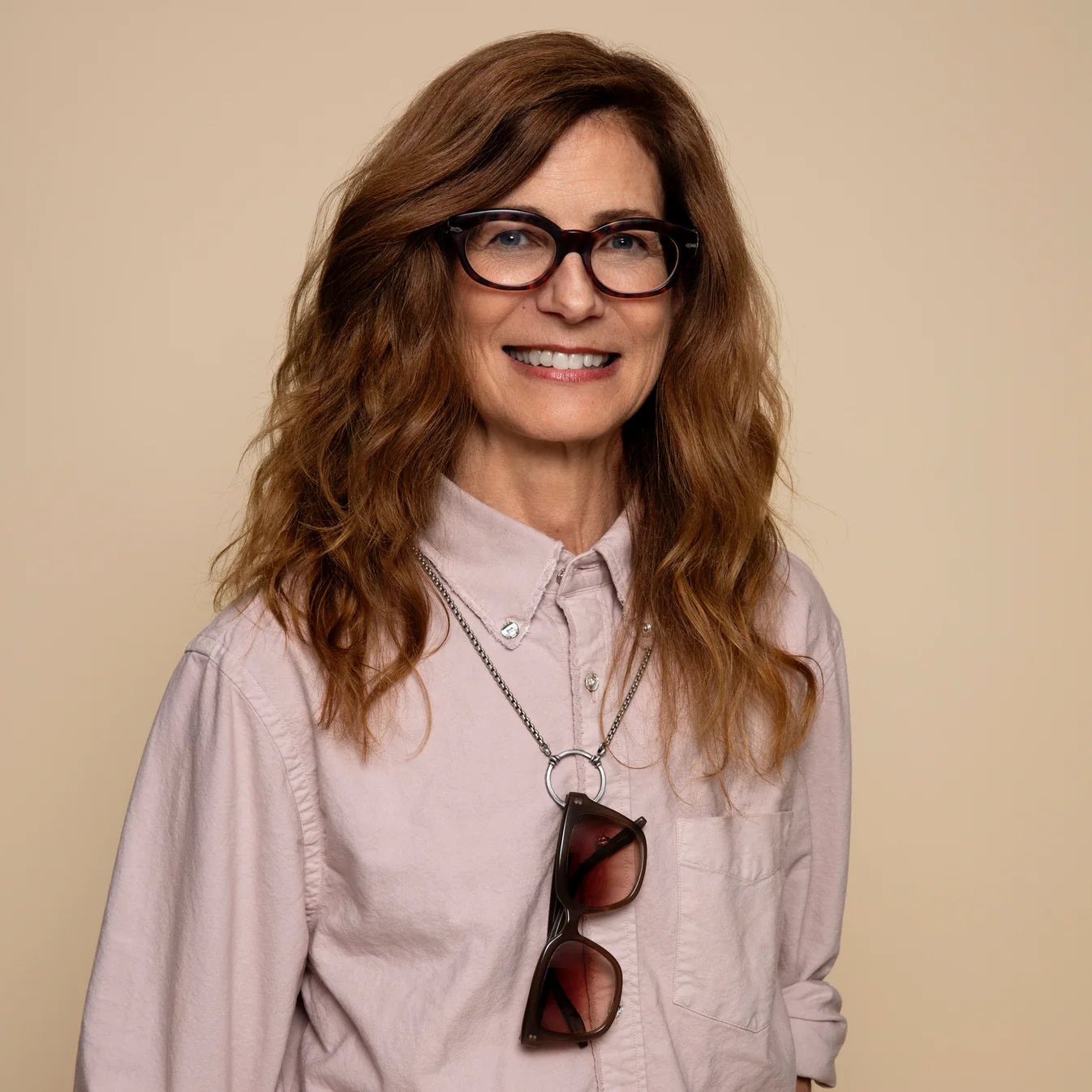 Woman wearing glasses and a light-colored shirt with sunglasses hanging from her neckline against a beige background