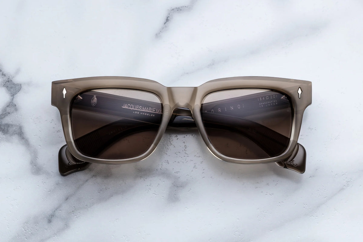 Jacques Marie Mage Torino translucent taupe rectangular ’60s-inspired sunglasses with dark lenses displayed on a white marble surface.