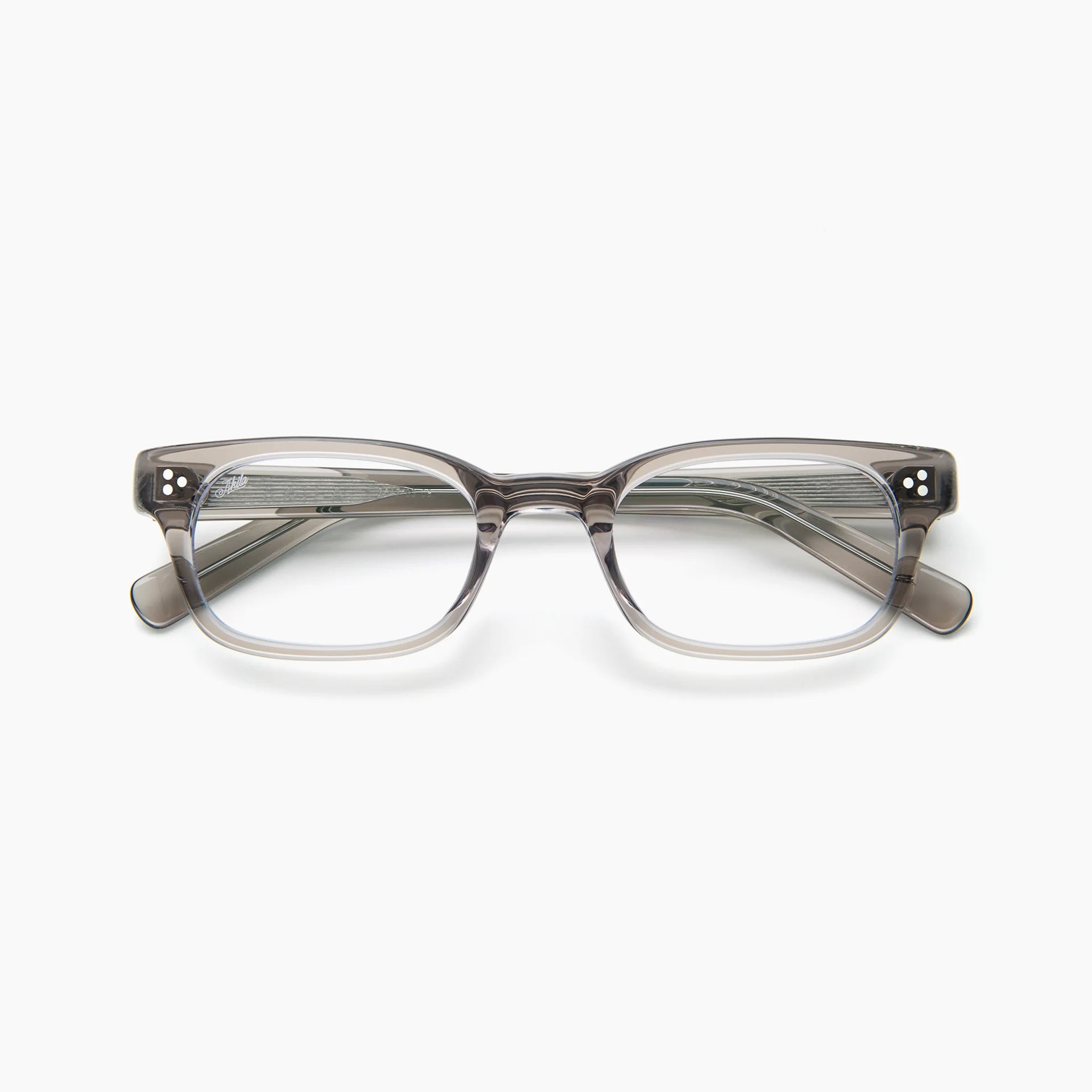The Akila Tracer is a gray rectangular eyeglass frame with clear lenses, displayed front-facing on a white background. This Akila design features arms that angle slightly outward and showcases small silver dot accents on the corners. The Tracer frame is also compatible with blue light blocking lenses for enhanced eye protection.