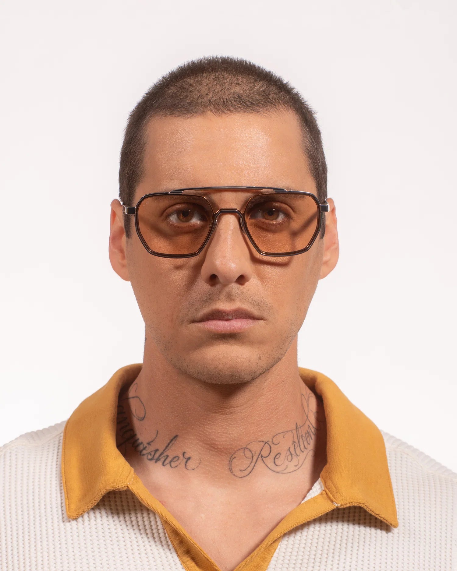 A man wearing thick, angular aviator-style sunglasses with amber-tinted lenses and dark brown frames, paired with a yellow polo shirt collar, against a plain white background.