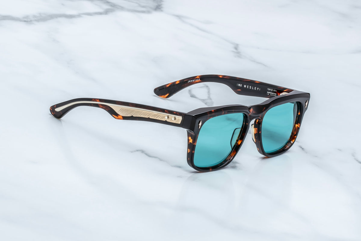 Inspired by the Old West, the Jacques Marie Mage Wesley sunglasses feature a striking tortoiseshell frame, teal blue lenses, and gold detailing on the arms, elegantly displayed atop a white marble surface—charismatic collectibles reimagined as modern rectangular spectacles by Jacques Marie Mage.