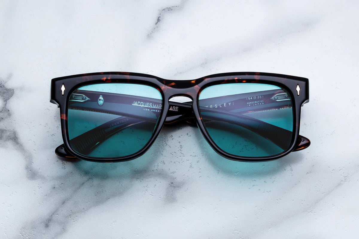 Jacques Marie Mage Wesley sunglasses featuring a square tortoiseshell frame and blue-tinted lenses rest on a white marble surface, their reflective finish and Jacques Marie Mage branding inside the arms evoking Old West-inspired rectangular spectacles with charismatic collectible appeal.