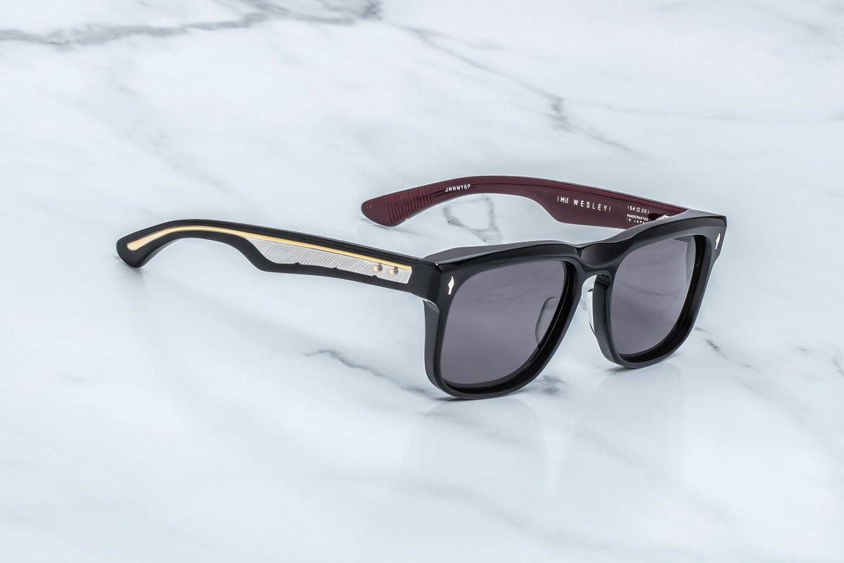 The Jacques Marie Mage Wesley sunglasses by Jacques Marie Mage are striking black rectangular frames featuring gold detailing on the temples, displayed atop a white marble surface. The dark red inner arms with white text lend these collectible spectacles an Old West-inspired flair.