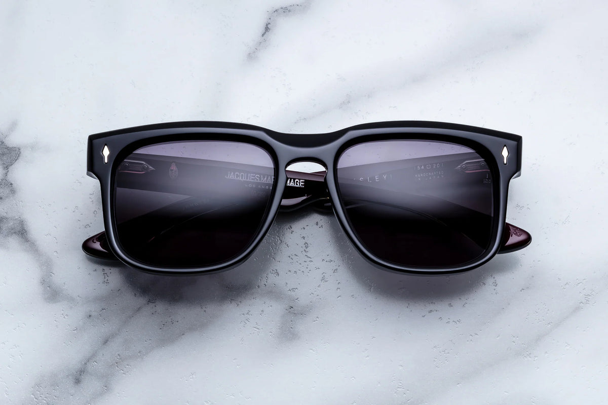 Jacques Marie Mage Wesley rectangular sunglasses with dark lenses rest on a white marble surface. These Jacques Marie Mage frames feature silver corner details, adding an Old West-inspired accent to their modern design.