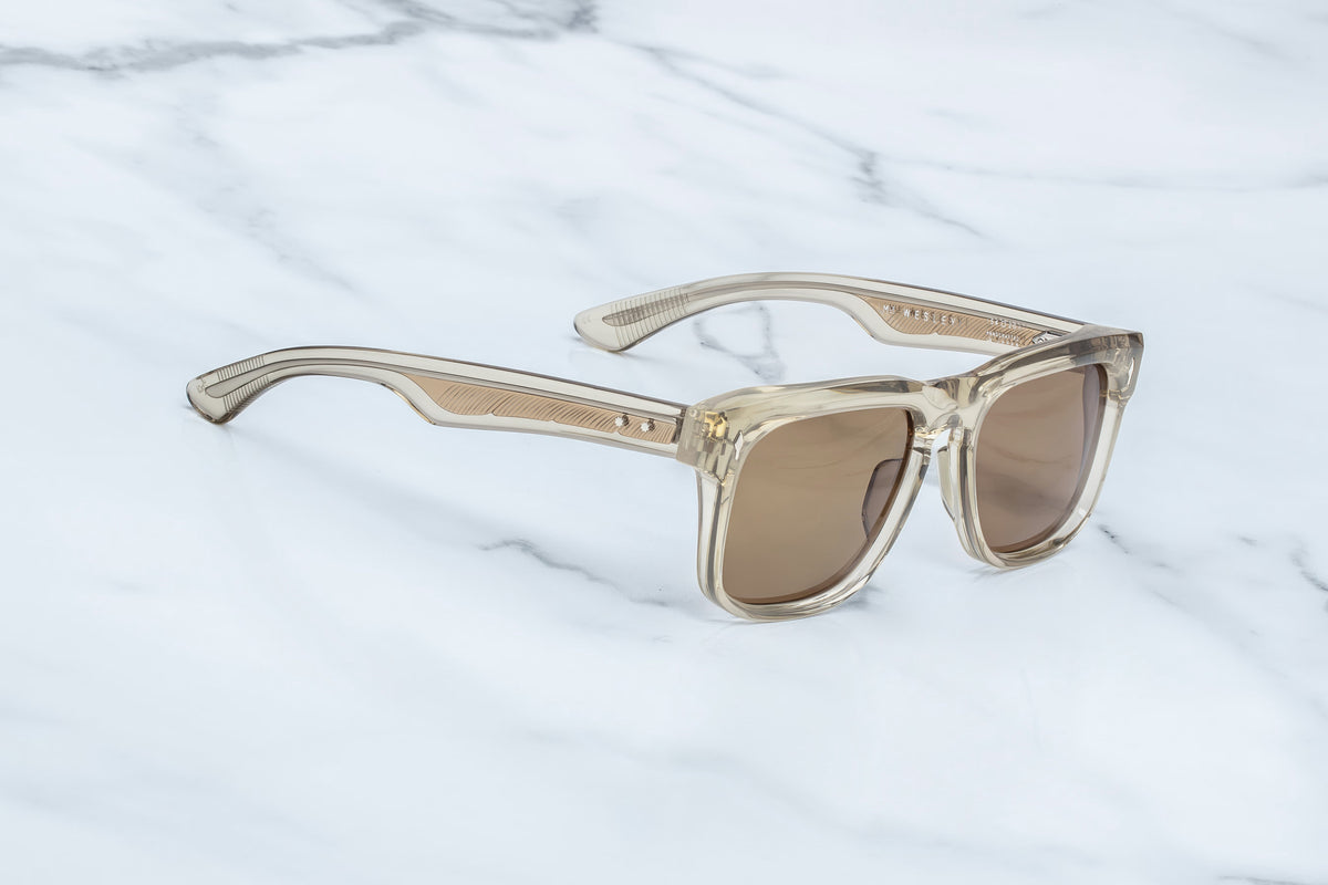 The Jacques Marie Mage Wesley sunglasses by Jacques Marie Mage, featuring transparent beige thick frames and brown-tinted lenses reminiscent of feathered wirecore spectacles, are placed on a white marble surface.