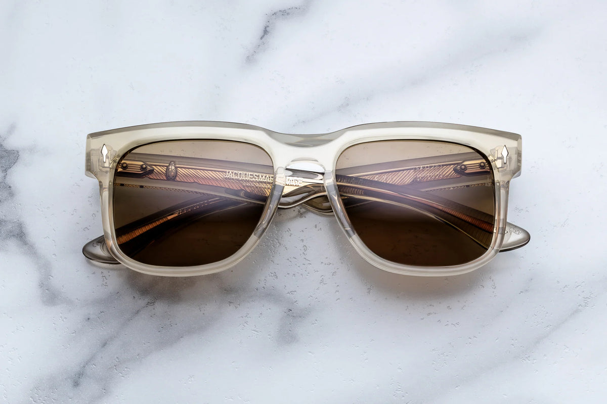 The Jacques Marie Mage Wesley sunglasses by Jacques Marie Mage, featuring clear frames and brown-tinted lenses that evoke Old West-inspired feathered wirecore spectacles, are displayed on a white marble surface with subtle gray veining. The arms of these charismatic collectibles are crossed behind the lenses.