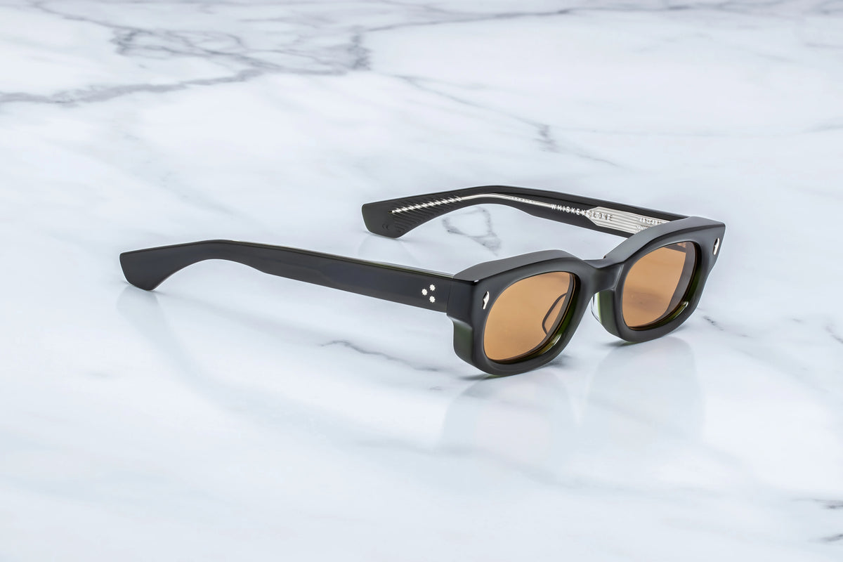 Jacques Marie Mage Whiskeyclone black-framed 90s rectangular sunglasses with amber-tinted lenses resting on a marble surface, radiating a sense of sartorial cynicism.