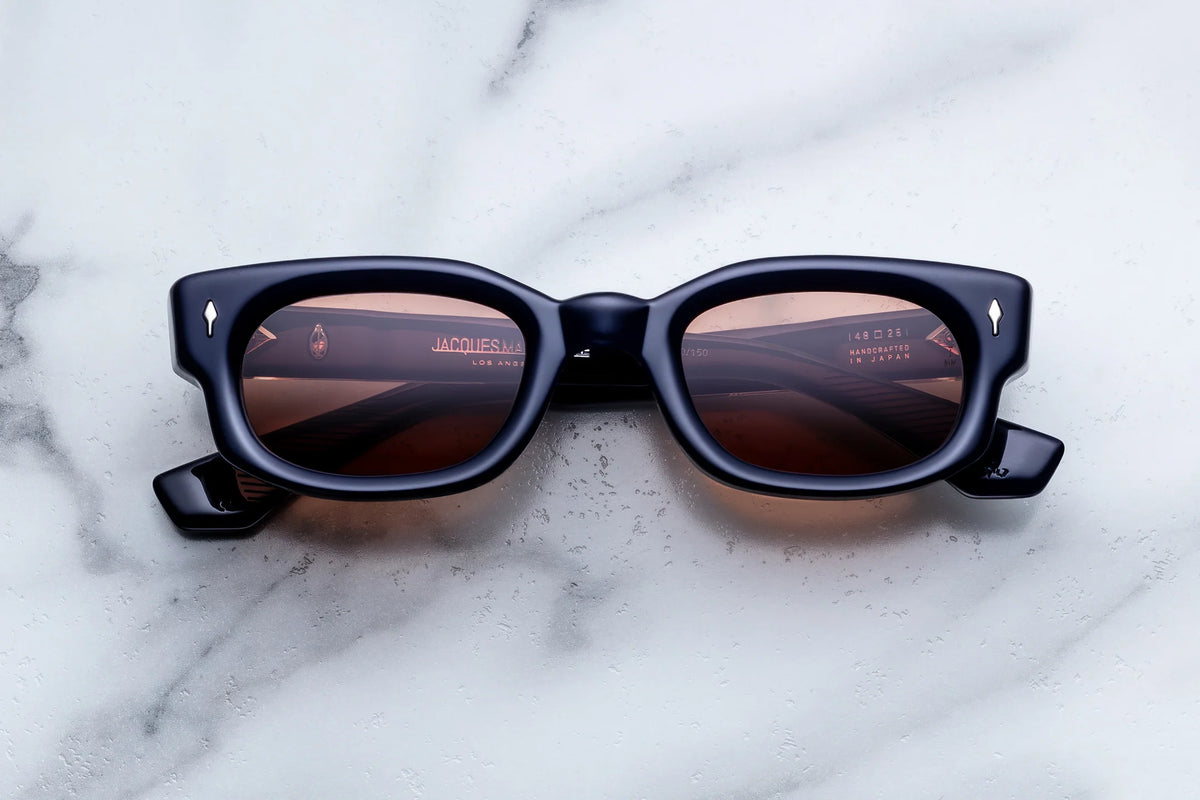 Jacques Marie Mage Whiskeyclone sunglasses with dark rims and reflective lenses resting on a marble surface.