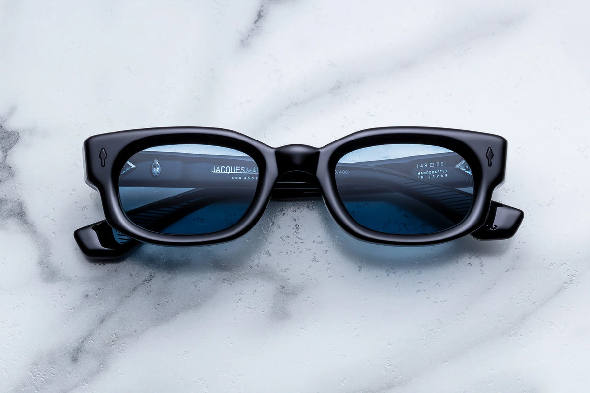 Jacques Marie Mage Whiskeyclone black rectangular-frame sunglasses with blue-tinted lenses and a sense of sartorial cynicism rest on a marble surface.