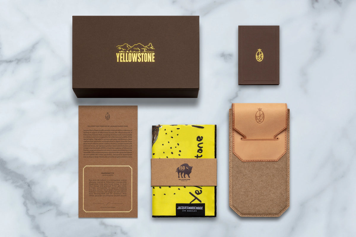 Jacques Marie Mage Yellowstone VII: Barronette In Brush shown with a brown presentation box, a small card, an informational pamphlet, a yellow patterned cloth, and a tan leather carrying case for the handcrafted polarized mineral glass eyewear.