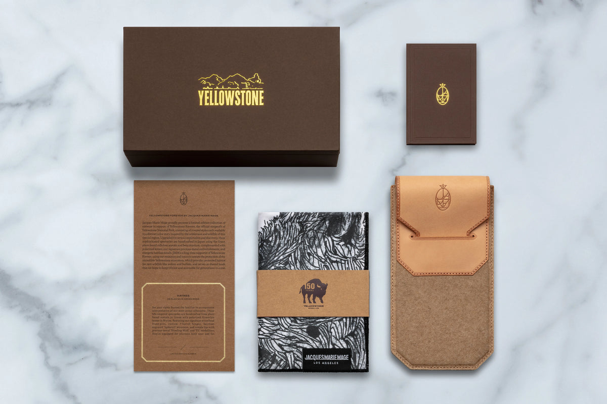 Jacques Marie Mage Yellowstone VII: Haynes In Green Moss gift set including a large brown storage box, a small card, an information sheet, a patterned microfiber cloth, and a tan leather case for ’60s-inspired spectacles on a marble surface.