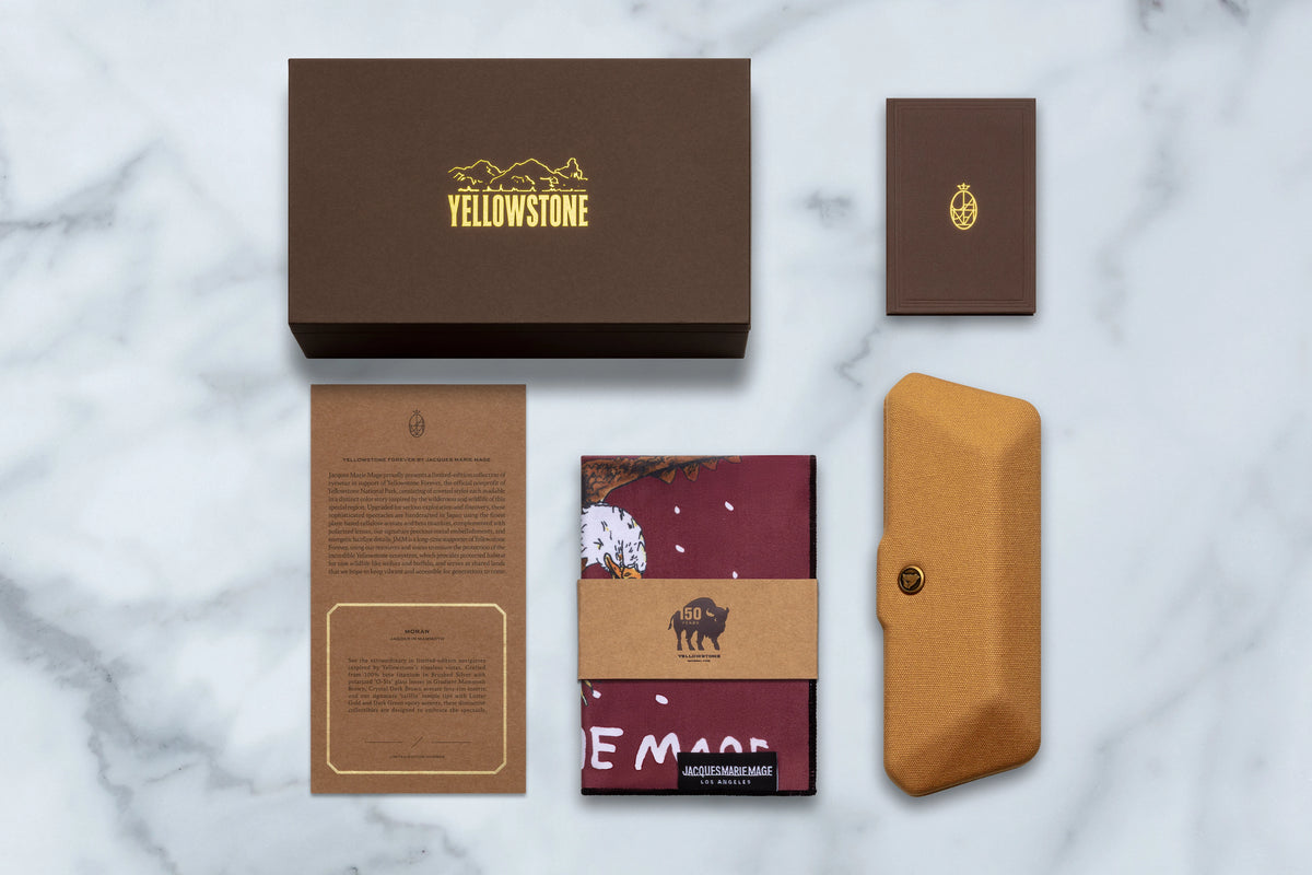 A brown storage box, a small card, a descriptive pamphlet, a patterned cleaning cloth, and a tan hard-shell case for the Jacques Marie Mage Yellowstone VII: Moran In Mammoth sunglasses arranged on a white marble surface.