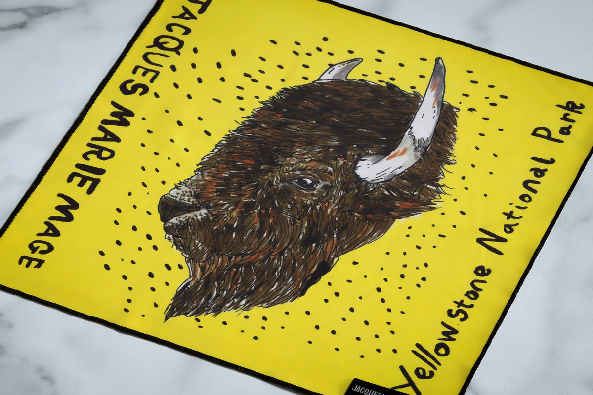 A yellow square microfiber cloth for Jacques Marie Mage Yellowstone VII: Barronette In Brush eyewear, featuring a bison head illustration surrounded by dots and brand lettering.