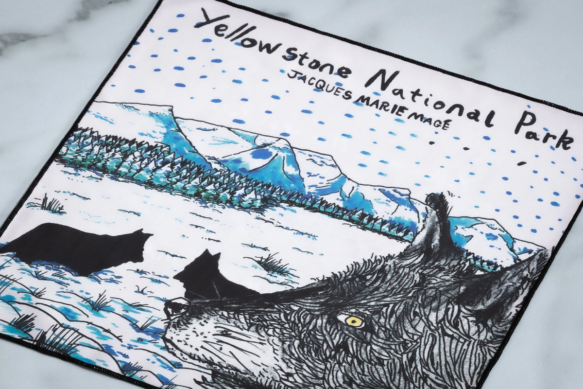 A white microfiber cleaning cloth for Jacques Marie Mage Yellowstone VII: Haynes In Green Moss featuring an illustrated winter scene of a wolf, snow-covered mountains, and the text Yellowstone National Park.