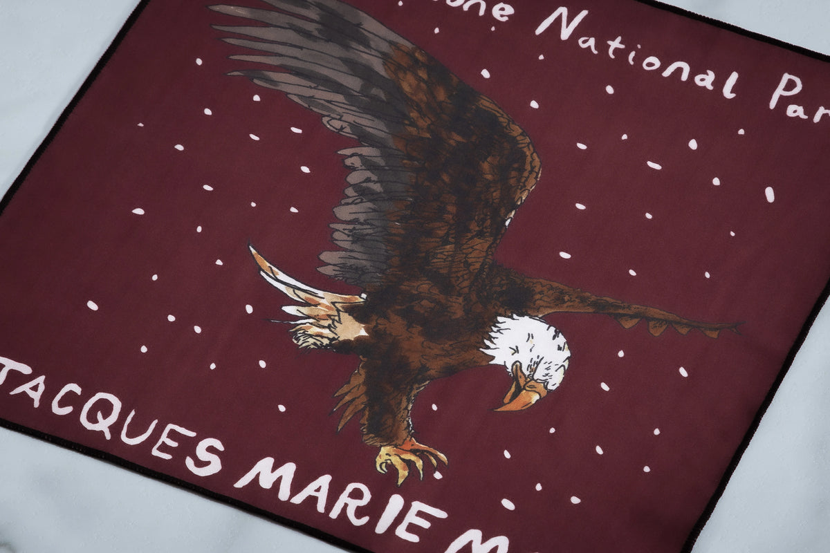A maroon cloth from the Jacques Marie Mage Yellowstone VII: Moran In Mammoth collection featuring a bald eagle illustration, white speckles, and text reading Yellowstone National Park and Jacques Marie Mage.