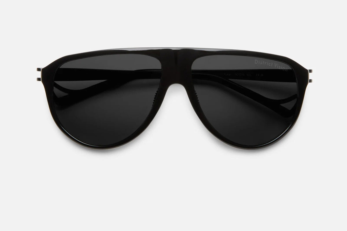 District Vision Yukari Windshield sunglasses by District Vision, shown from the front against a plain light gray background, feature a classic black aviator silhouette with dark lenses. The sleek six-base frame offers enhanced durability while evoking a stylish windshield-inspired design.