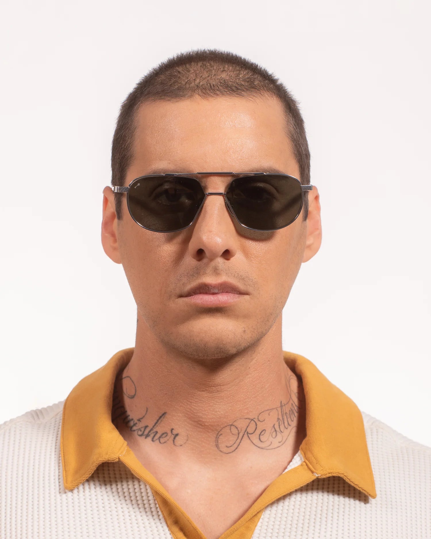 A person with short hair, visible neck script tattoos, and a beige shirt with a yellow collar faces the camera while wearing luxury Leisure Society Yuma sunglasses in dark gold ion plating. The background is plain white.