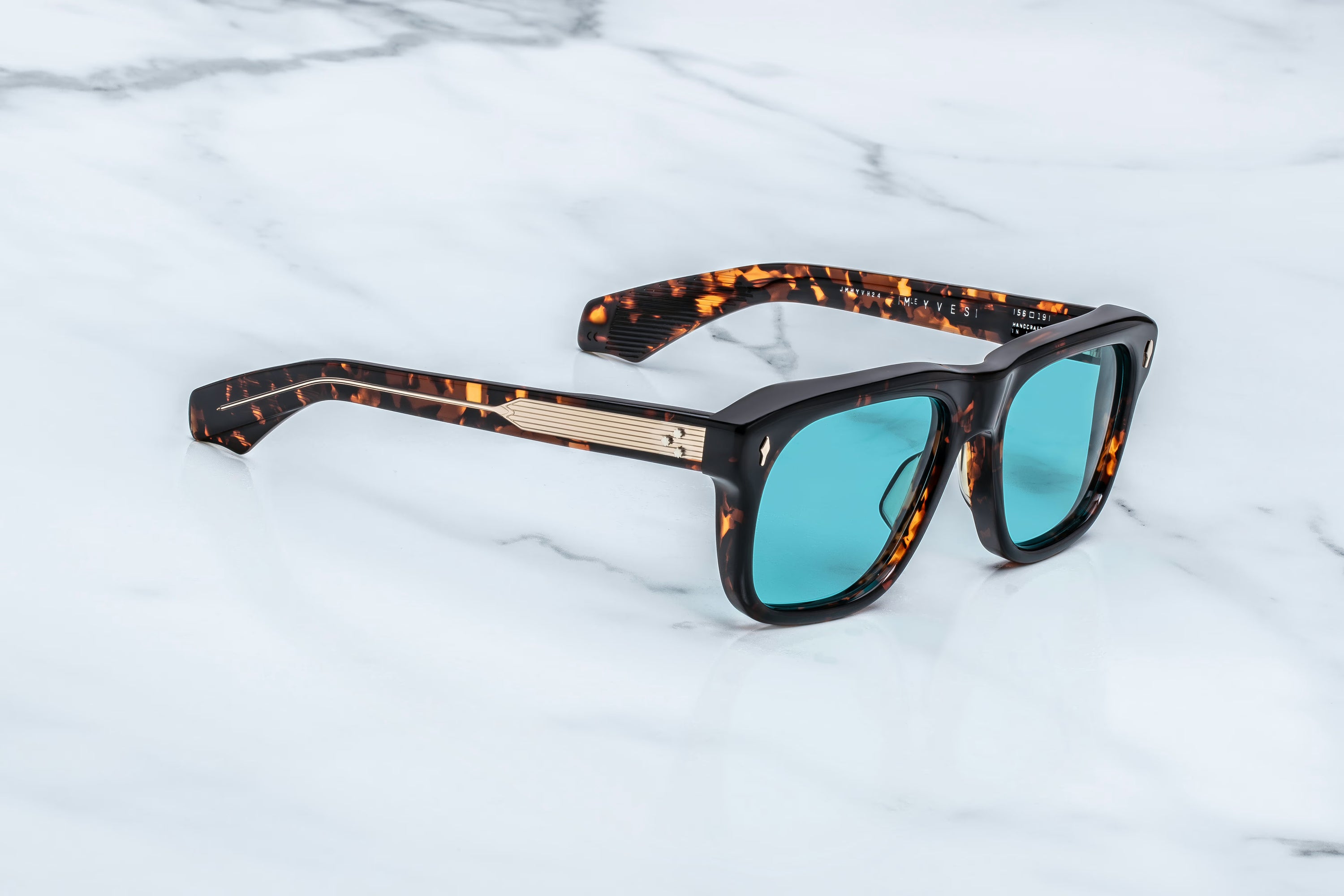 Jacques Marie Mage Yves sunglasses from Jacques Marie Mage, featuring a 70’s-inspired tortoiseshell frame and teal lenses, rest on a white marble surface, evoking the elegance of French couturiers.