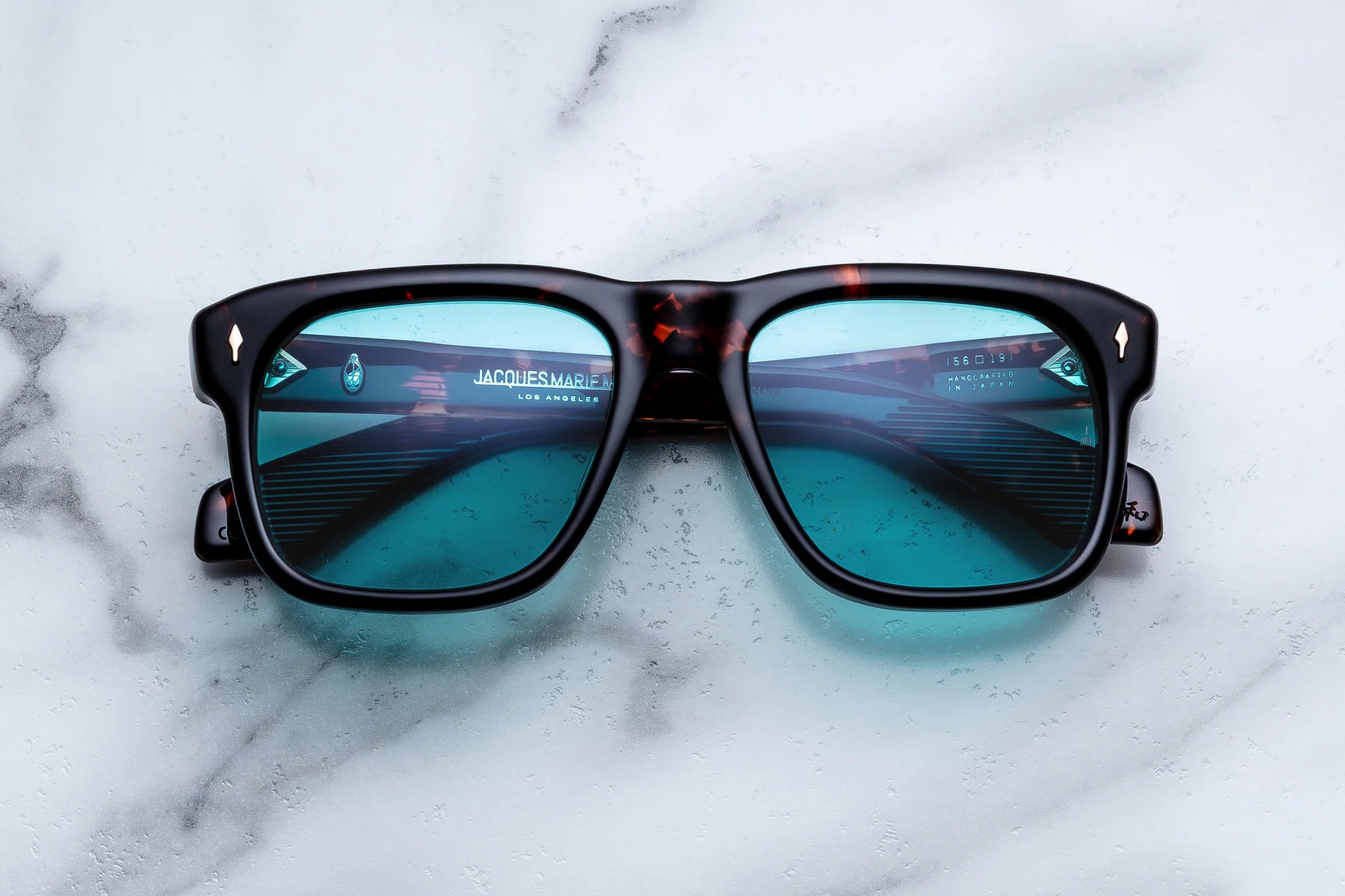 The Jacques Marie Mage Yves sunglasses by Jacques Marie Mage feature a 70s-inspired tortoiseshell design with teal-tinted lenses, elegantly displayed on a white marble surface. Each limited-edition pair reflects a modern architectural scene in the lenses, evoking the effortless sophistication of French couturiers while merging retro flair with contemporary style.