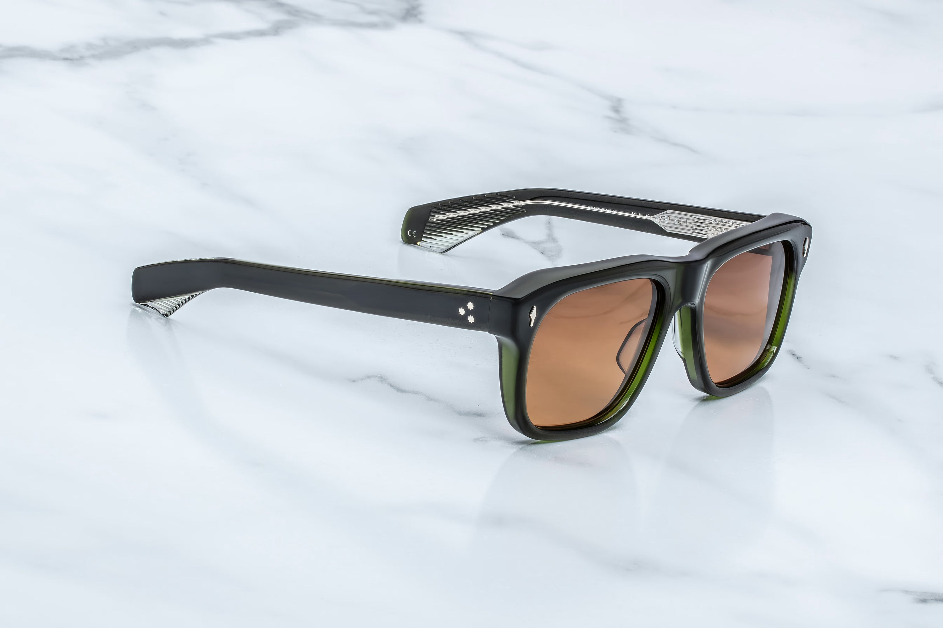 Square-framed sunglasses with a translucent black front and green-tinted acetate arms, featuring warm brown-tinted lenses, displayed on a white marble surface.