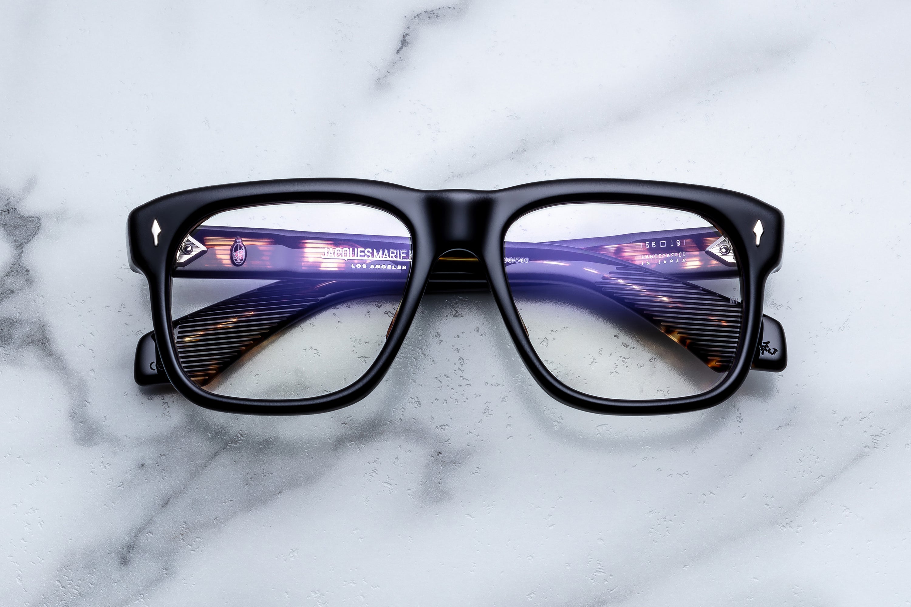 Jacques Marie Mage Yves black rectangular eyeglasses with thick, iconic frames rest on a white marble surface, their lenses reflecting text and surroundings—a style inspired by the legendary fashion designer and namesake.