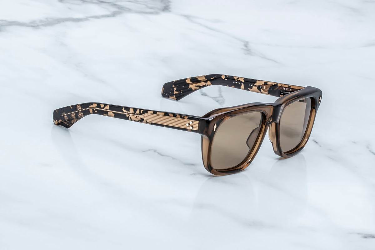 Jacques Marie Mage Yves sunglasses by Jacques Marie Mage, featuring a bold brown square frame, dark lenses, and intricately patterned arms, rest on a white marble surface—exuding a 70s-inspired flair reminiscent of French couturiers.