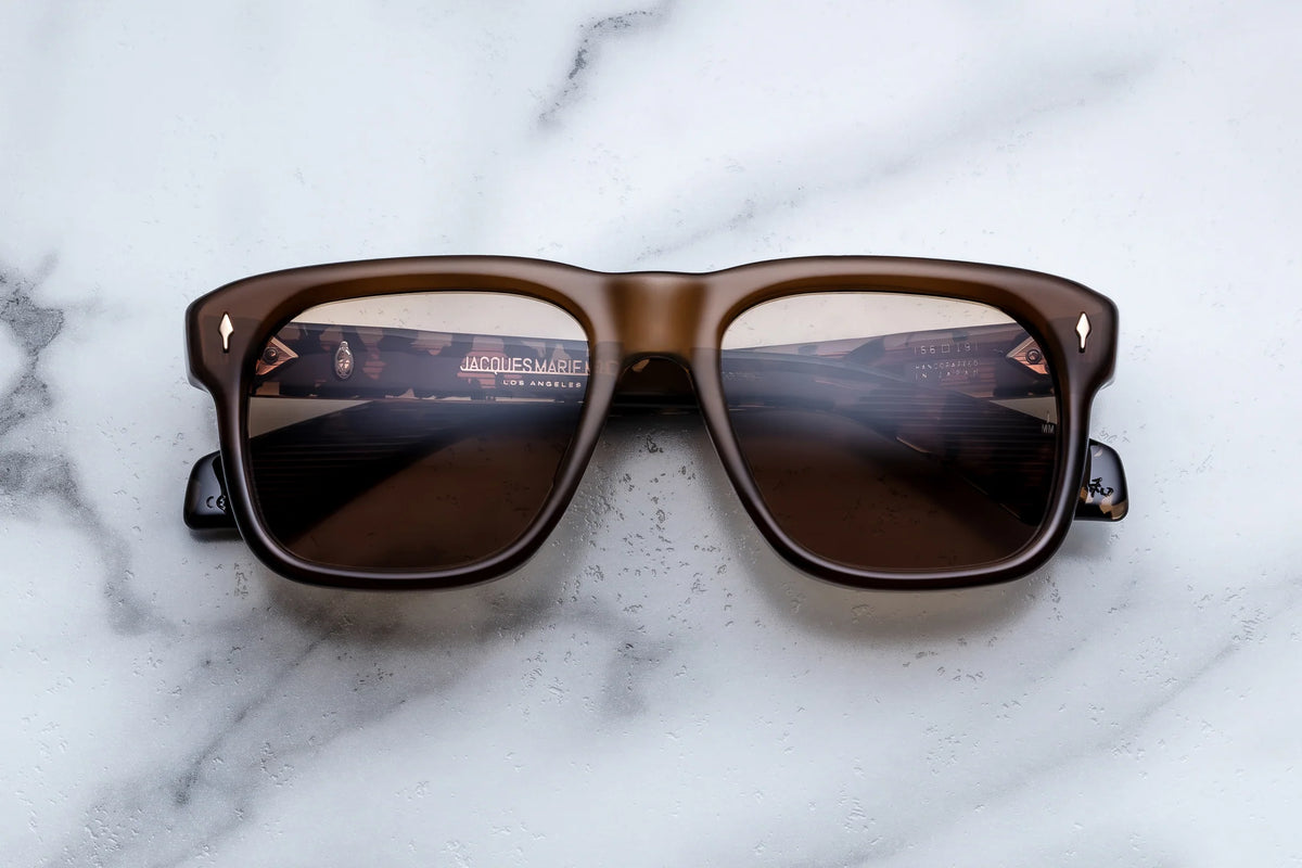 The Jacques Marie Mage Yves sunglasses, featuring thick brown frames inspired by iconic 70’s style revered by French couturiers, are displayed on a white marble surface. Their lenses reflect an indoor scene illuminated with lights and signs, suggesting the ambiance of a store or shopping area.