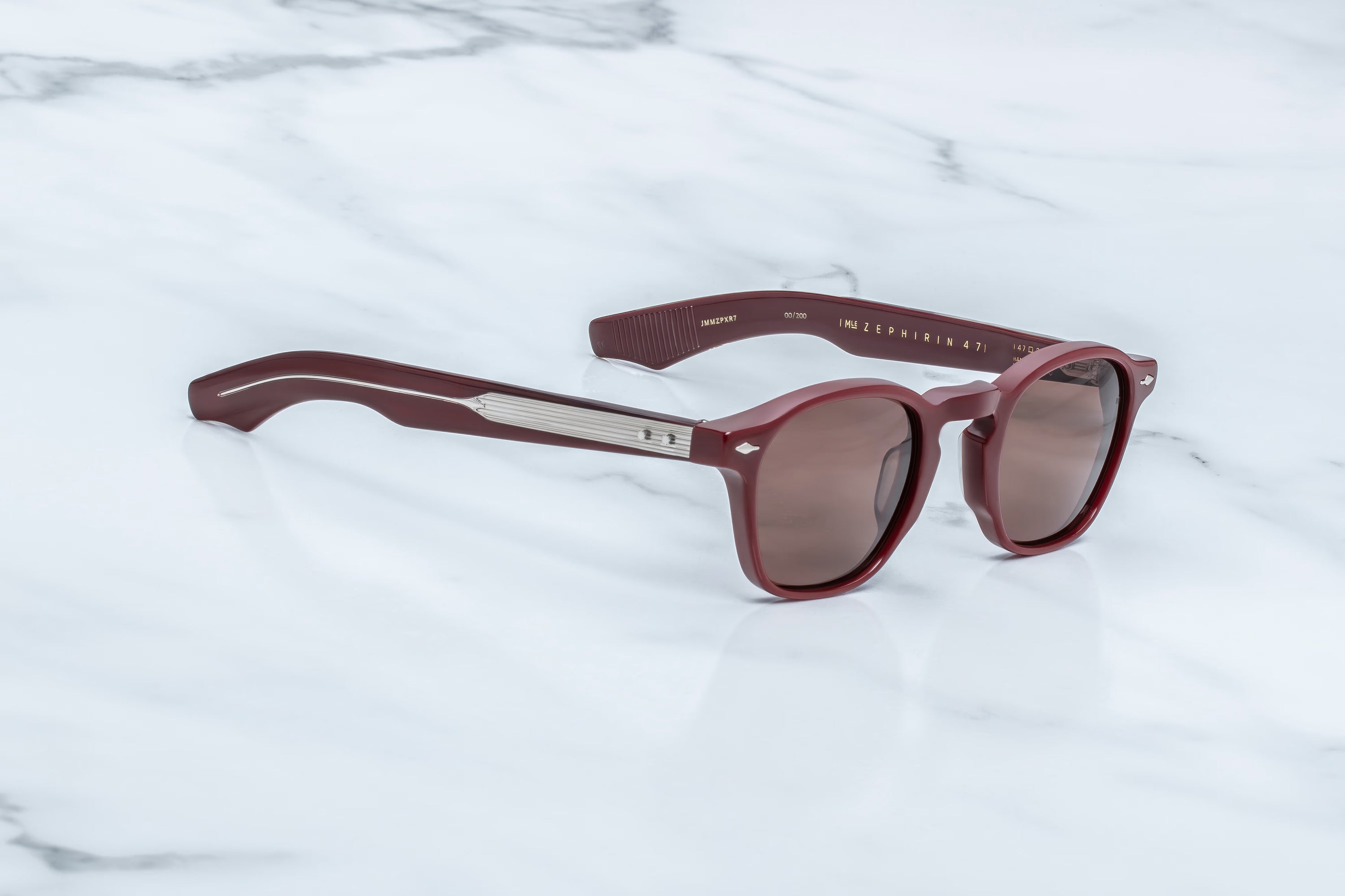 A pair of round sunglasses with a translucent burgundy frame and brown tinted lenses, resting on a white marble surface. The brand name 'MILANO' is printed on the temple arm.