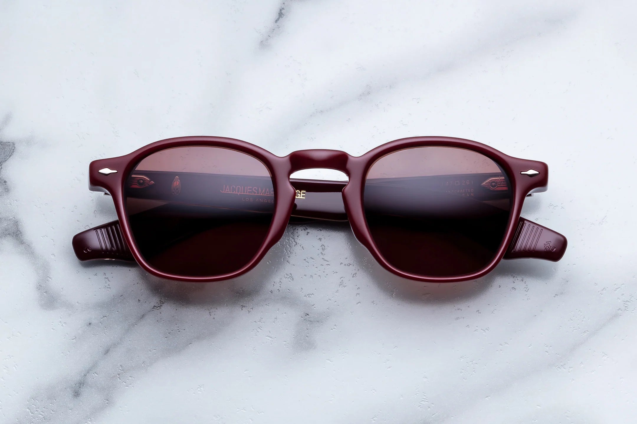 A pair of round-frame sunglasses with a translucent burgundy frame and dark tinted lenses, resting on a white marble surface.