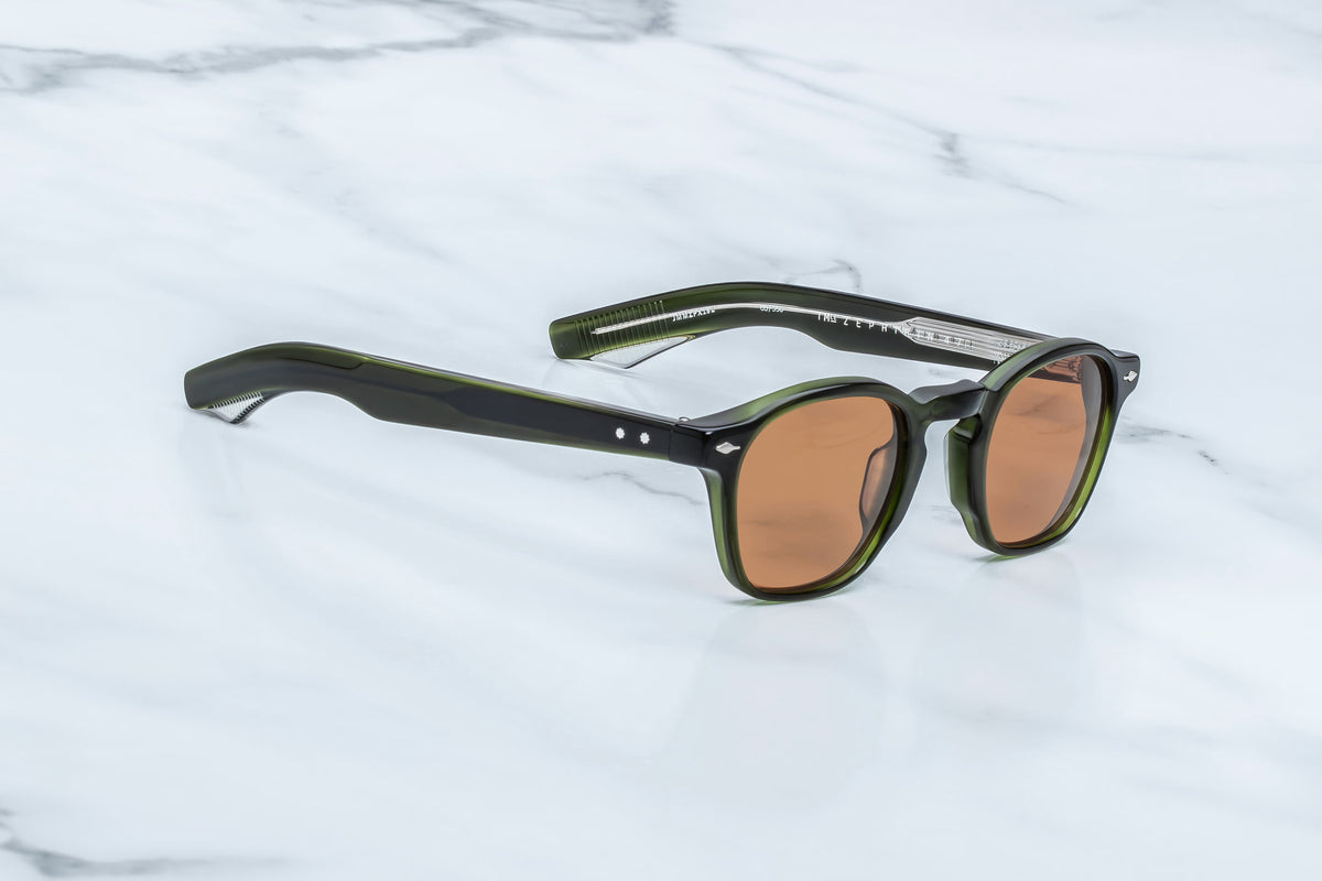 Jacques Marie Mage Zephirin 47 spectacles by Jacques Marie Mage, featuring an olive green and black hand-finished frame with brown lenses, rest on a white marble surface.