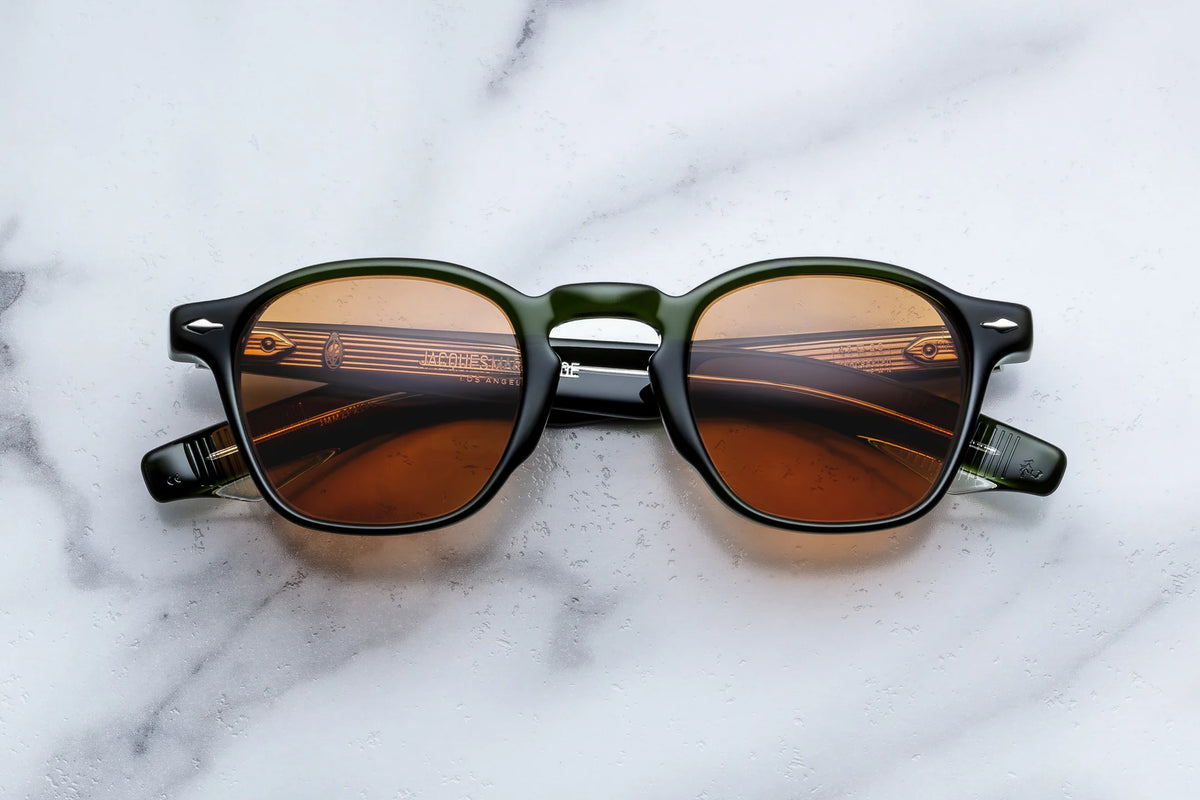 The Jacques Marie Mage Zephirin 47 by Jacques Marie Mage, hand-finished round dark olive sunglasses with brown lenses, rest on a white marble surface. These glasses showcase temples adorned with a subtle striped pattern and metallic accents near the hinges, capturing the timeless charm of vintage spectacles.