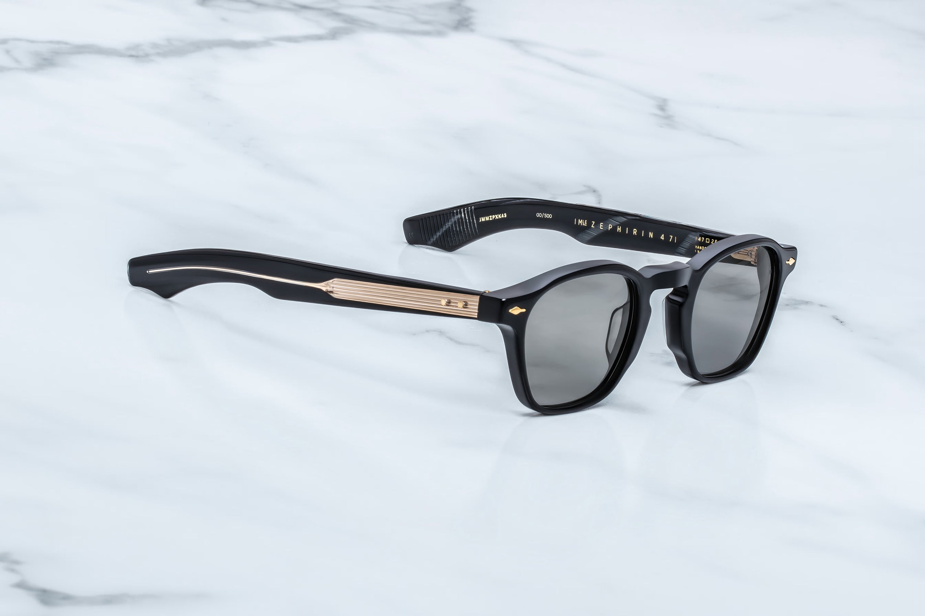 Black acetate sunglasses with a distinctive gold metallic accent on the bridge and temples, resting on a white marble surface.