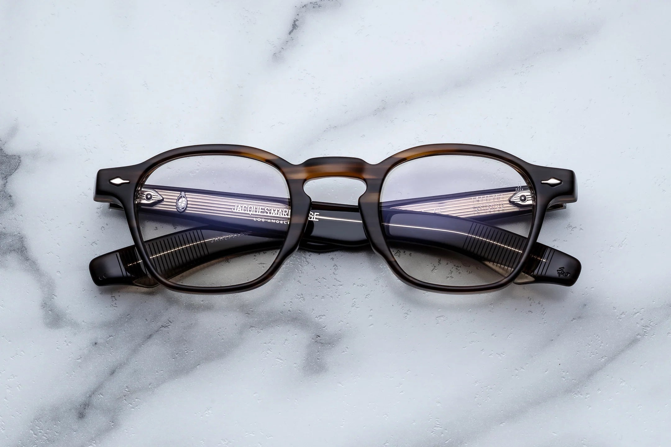 Jacques Marie Mage optical glasses in dark tortoise acetate with clear lenses and translucent striped temple tips, shown from the front on a light marble surface.