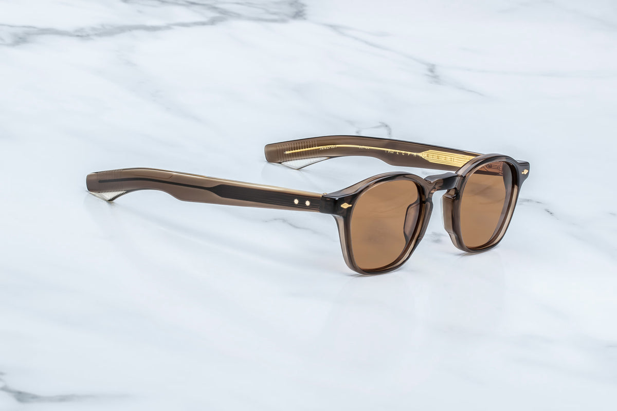 Jacques Marie Mage Zephirin 47 sunglasses with round, brown-tinted lenses rest on a white marble surface. Featuring thick, glossy frames and temples hand-finished to evoke pre-WWII sizing, these sunglasses by Jacques Marie Mage are reminiscent of classic ZEPHIRIN spectacles.