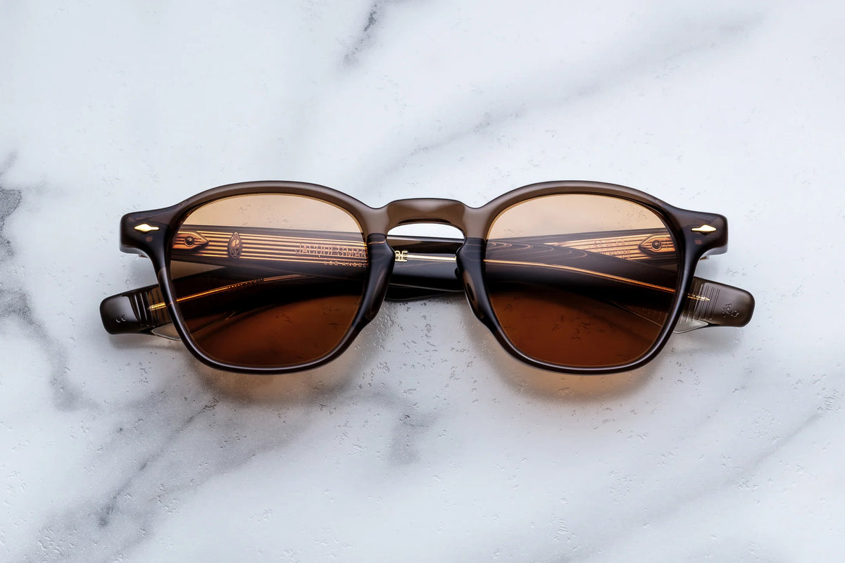 Jacques Marie Mage Zephirin 47 sunglasses by Jacques Marie Mage—featuring round brown frames with dark lenses—are placed upside down on a white marble surface. Inspired by pre-WWII sizing, the design showcases subtle gold accents on the arms, echoing the classic hand-finished craftsmanship of traditional spectacles.