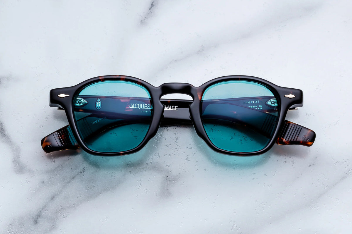 Jacques Marie Mage Zephirin spectacles in tortoiseshell-patterned acetate with teal lenses and hand-finished metal temple hinges, resting on a white marble surface.