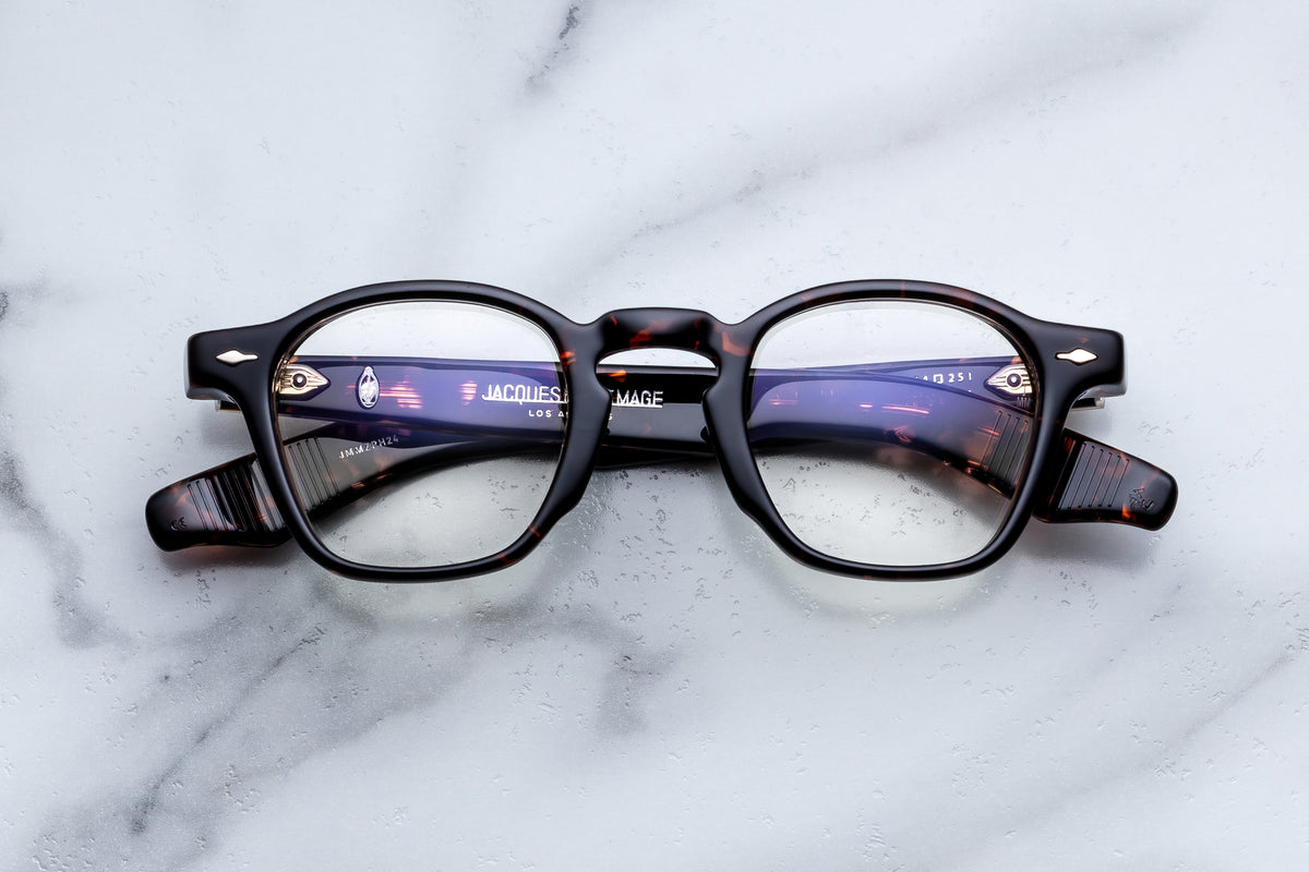 Jacques Marie Mage Zephirin hand-finished tortoiseshell acetate eyeglasses with a rounded frame and gold-toned metal detailing at the hinges, resting on a white marble surface.
