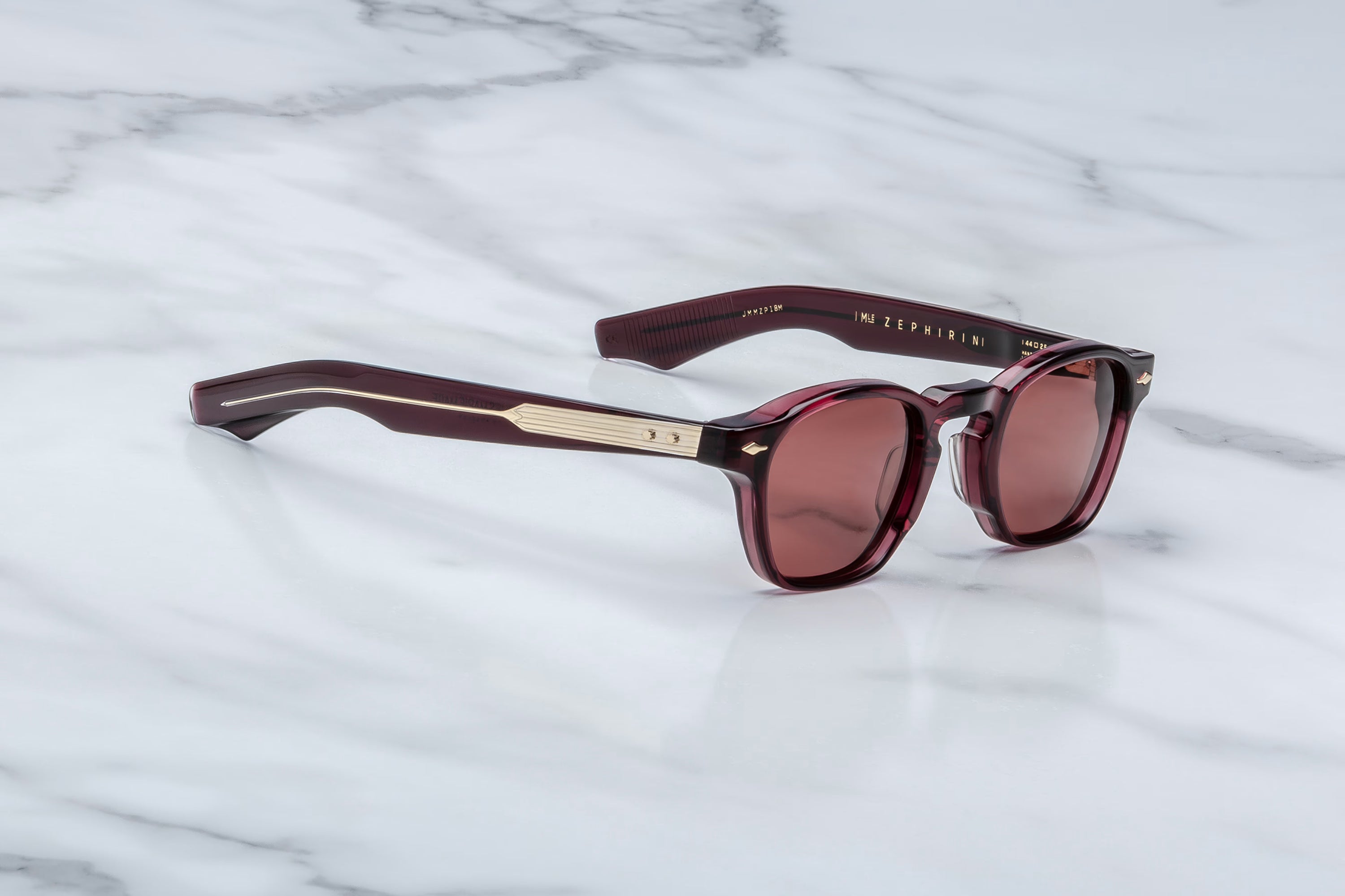 Jacques Marie Mage sunglasses in deep burgundy acetate with burgundy tinted lenses and gold-tone temple detailing, shown at an angle on a light marble surface.