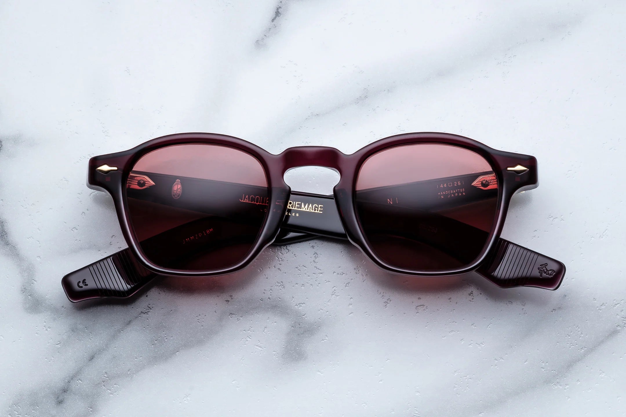 Jacques Marie Mage sunglasses in deep burgundy acetate with burgundy tinted lenses and translucent striped temple tips, shown from the front on a light marble surface.