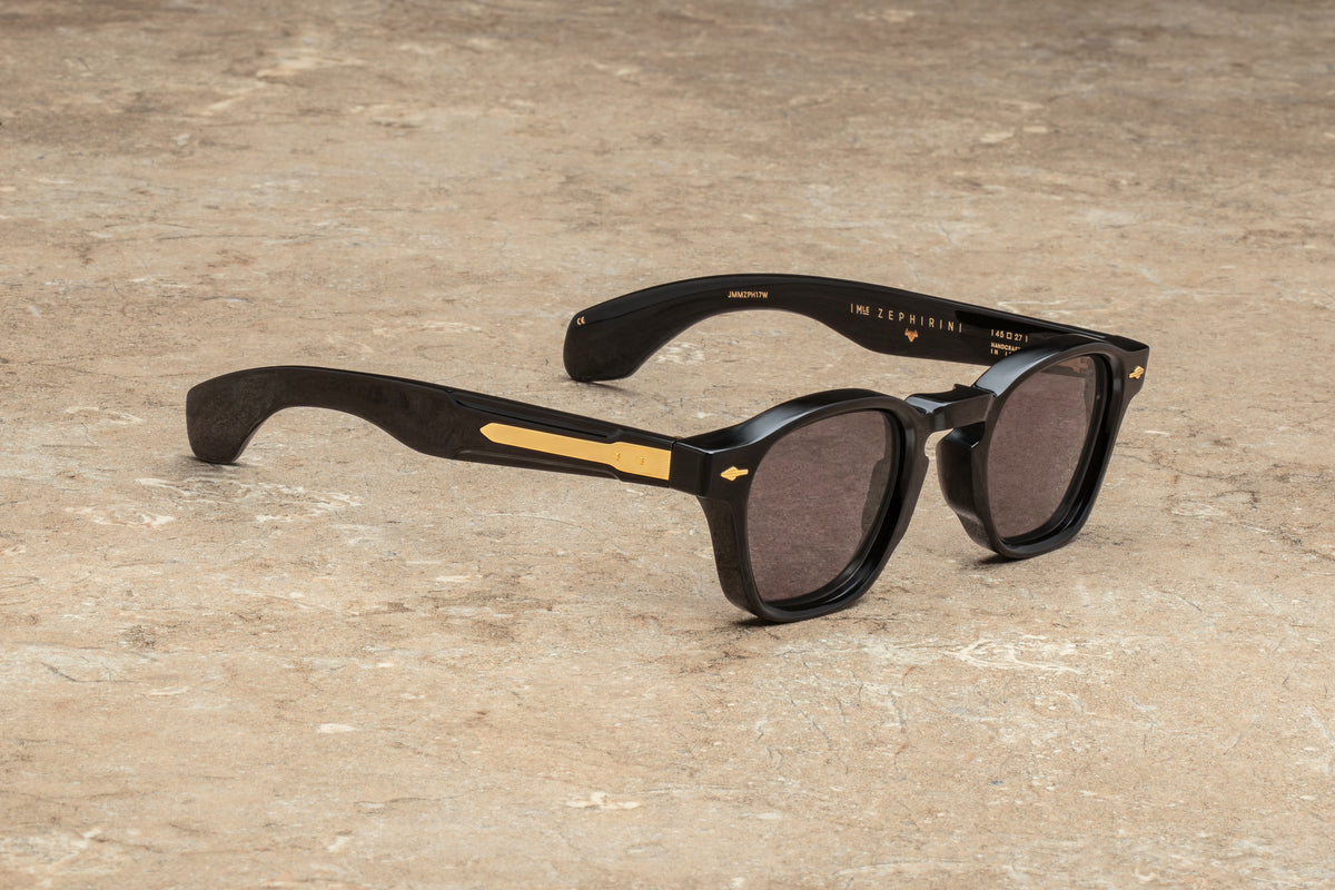 The Jacques Marie Mage Heritage 2: Zephirin sunglasses feature a bold black square frame with refined gold accents on the arms, resting elegantly on a textured stone surface. Their dark lenses and the brand’s gold-printed name on the inside of the right arm highlight the sophistication of handcrafted Italian eyewear, subtly echoing Art Deco influences.