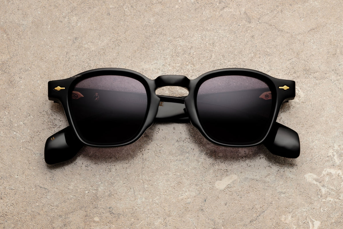 The Jacques Marie Mage Heritage 2: Zephirin sunglasses by Jacques Marie Mage feature bold black thick frames and dark lenses, resting on a light tan textured surface. Drawing inspiration from Art Deco eyewear, these sunglasses are adorned with subtle gold accents near the hinges, lending a vintage sophistication to their striking design.