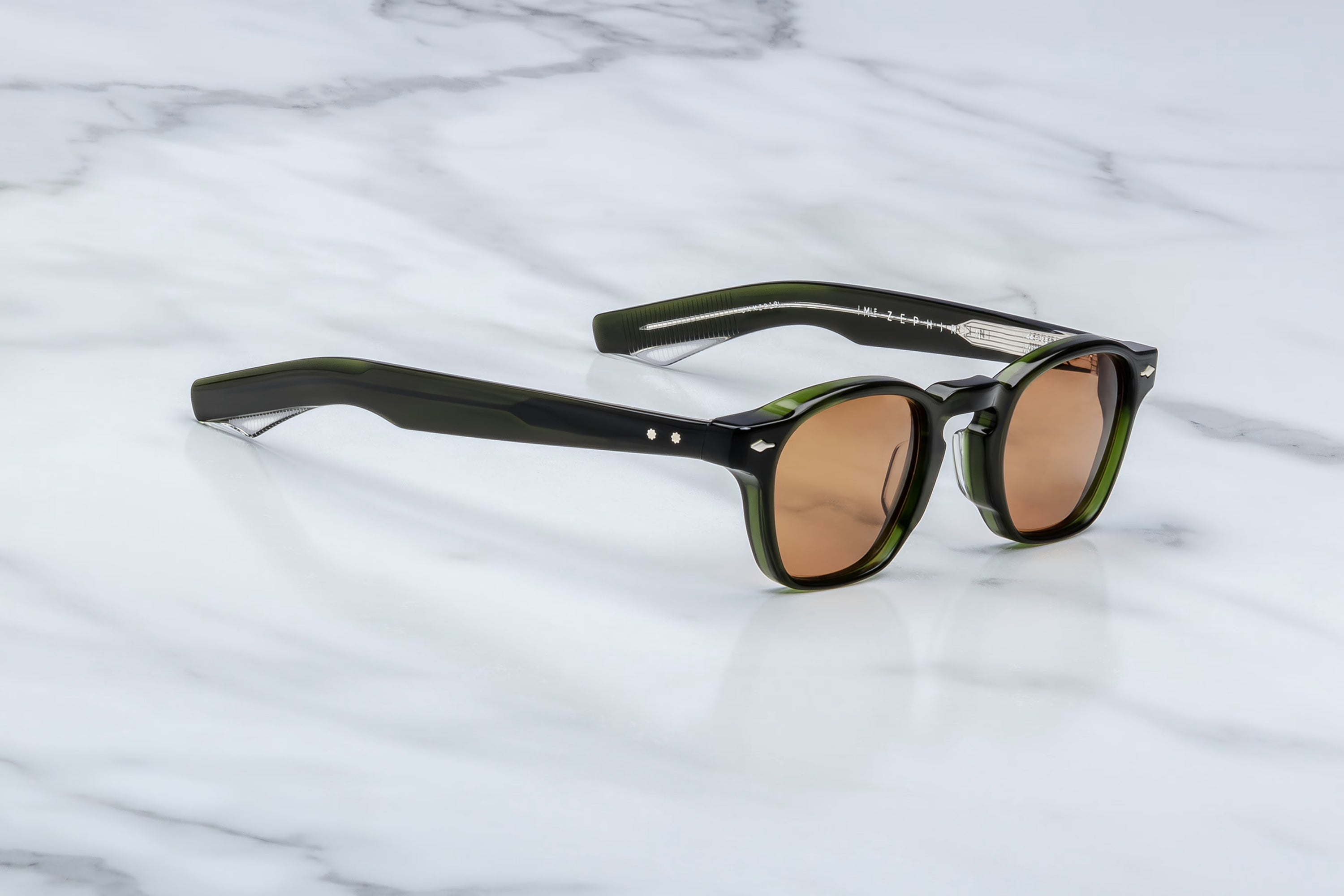 Jacques Marie Mage sunglasses in dark translucent green acetate with brown tinted lenses and clear striped temple tips, shown at an angle on a light marble surface.