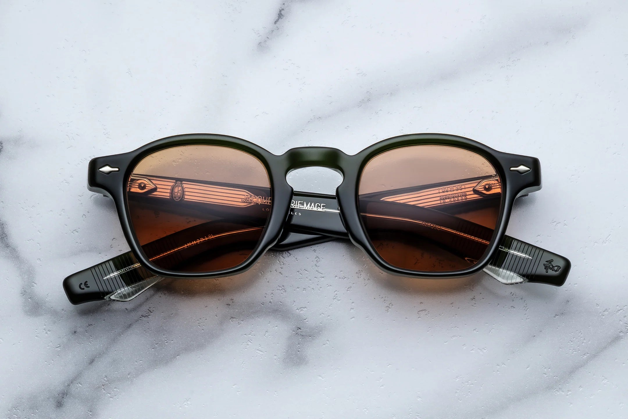 Jacques Marie Mage sunglasses in dark green acetate with brown tinted lenses and translucent striped temple tips, shown from the front on a light marble surface.