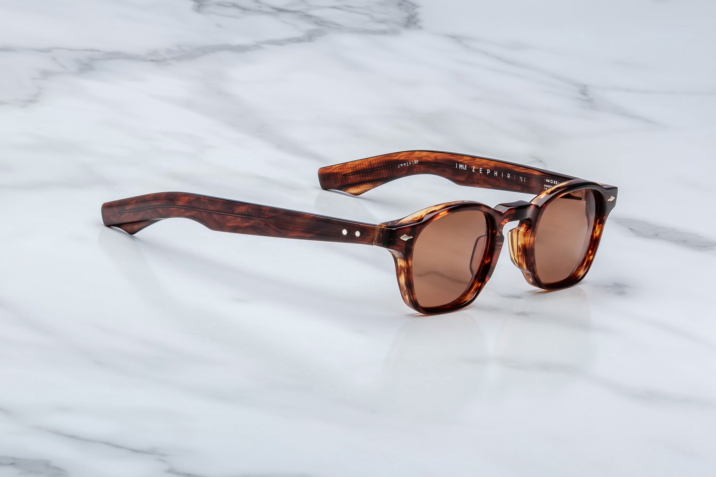 Jacques Marie Mage sunglasses in translucent brown acetate with brown tinted lenses, shown at an angle on a light marble surface.
