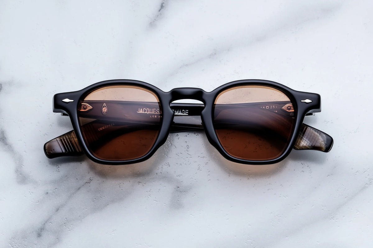 Black-framed Jacques Marie Mage Zephirin spectacles with brown-tinted lenses and sculpted temple construction resting on a marble surface.