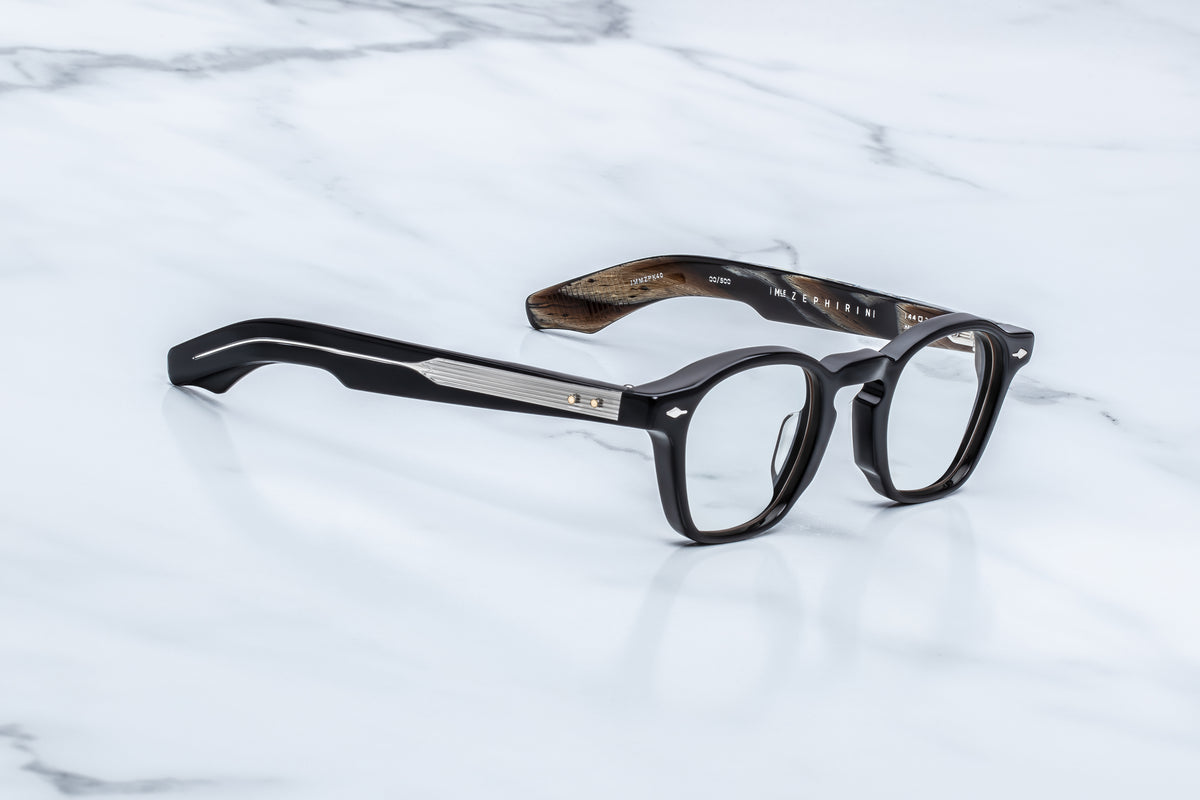 Black-rimmed Jacques Marie Mage Zephirin spectacles with metallic temples featuring silver-toned accents, resting on a white marble surface.